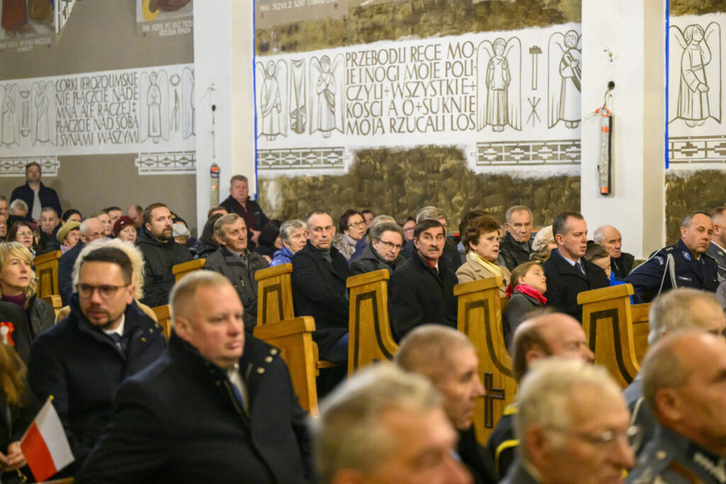 Na zdjęciu jest grupa ludzi w kościele. Wnętrze jest jasne, z drewnianymi ławkami i białymi ścianami z napisami. Osoby siedzą, niektóre trzymają flagi.