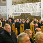 Na zdjęciu jest grupa ludzi w kościele. Wnętrze jest jasne, z drewnianymi ławkami i białymi ścianami z napisami. Osoby siedzą, niektóre trzymają flagi.