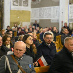 Ludzie siedzą w kościele, skupieni na wydarzeniu. Ubrani są w ciemne płaszcze, niektórzy trzymają polską flagę. W tle jasne ściany z dekoracjami.