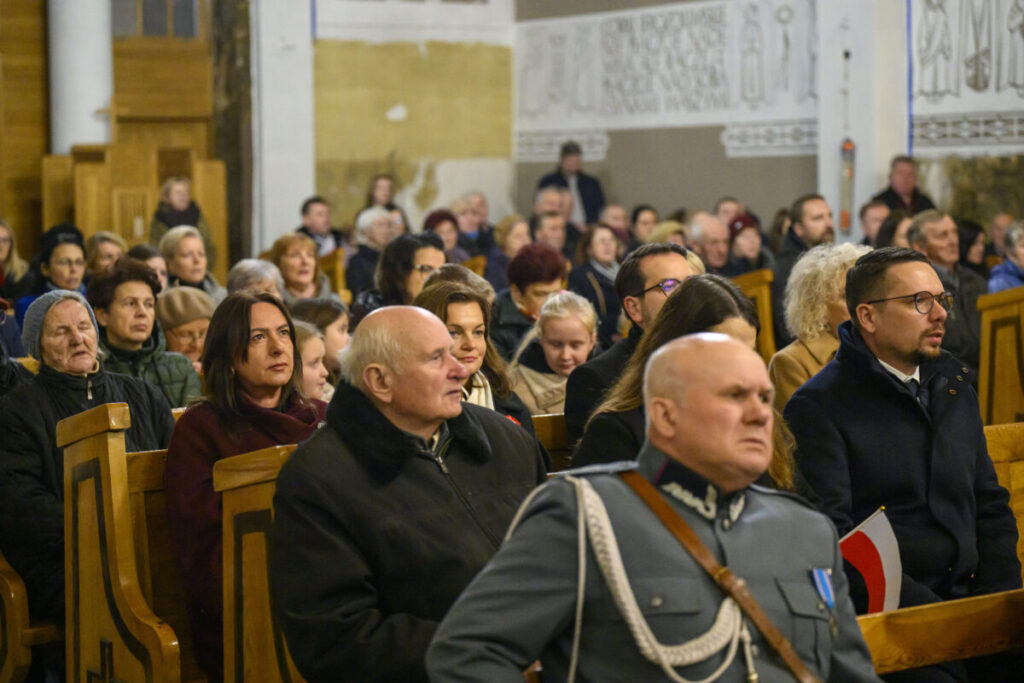 Na zdjęciu widać tłum ludzi siedzących w drewnianych ławkach w kościele. Dominują kolory ziemi i szarości, część osób ma na sobie ciepłe ubrania. Tło zdobią białe ściany z dekoracyjnymi napisami.