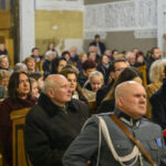 Na zdjęciu widać tłum ludzi siedzących w drewnianych ławkach w kościele. Dominują kolory ziemi i szarości, część osób ma na sobie ciepłe ubrania. Tło zdobią białe ściany z dekoracyjnymi napisami.