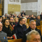 Zdjęcie ukazuje tłum ludzi w kościele, siedzących w drewnianych ławkach. Wiele z nich ma przypięte biało-czerwone kotyliony. Tło z jasnymi, zdobionymi ścianami.