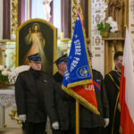 Grupa osób w mundurach stoi przed obrazem Jezusa Miłosiernego w kościele. Widoczne są flagi w kolorach czerwonym i niebieskim oraz białe kwiaty. Tło zdobią złote elementy i napisy na ścianie.