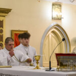 Wnętrze kościoła, kilku ministrantów w białych szatach przy ołtarzu. Złote i kremowe tło, jasne światło. Kapłan w złotej szacie, widać kielich.