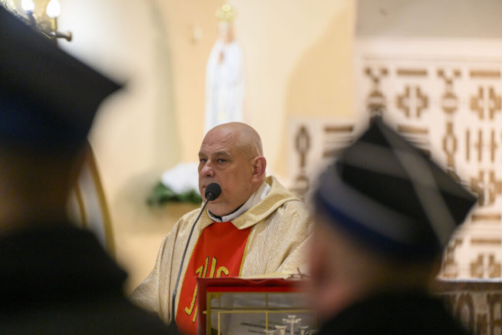 Mężczyzna w kościelnych szatach mówi do mikrofonu. Jasne tło z figurą świętą. Kolory szaty: złoty i czerwony.