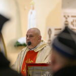 Mężczyzna w kościelnych szatach mówi do mikrofonu. Jasne tło z figurą świętą. Kolory szaty: złoty i czerwony.