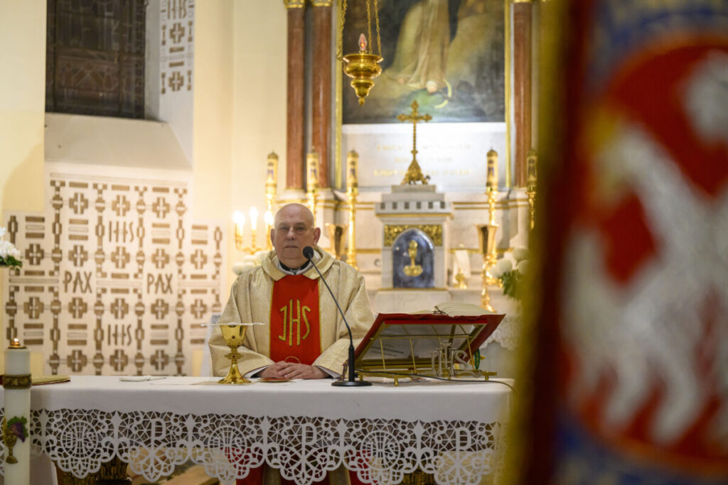 Kapłan w białych szatach stoi przy ołtarzu z mikrofonem. Tło ozdobione jest złotymi elementami i obrazem. Widać biały obrus z koronką.