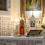 Kapłan w złotym ornacie stoi za ołtarzem, na którym znajduje się kielich. W tle obrazy religijne, świece i złote ozdoby. Wnętrze kościoła jest jasne i bogato zdobione.