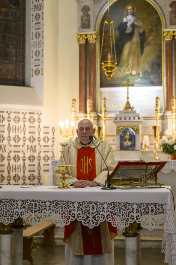 Kapłan w złotym ornacie stoi za ołtarzem, na którym znajduje się kielich. W tle obrazy religijne, świece i złote ozdoby. Wnętrze kościoła jest jasne i bogato zdobione.
