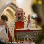 Na zdjęciu ksiądz w złotej szacie czyta przy ołtarzu. W tle widoczne są łuki i ściana w ciepłych barwach. Obok otwarta księga i mikrofon.