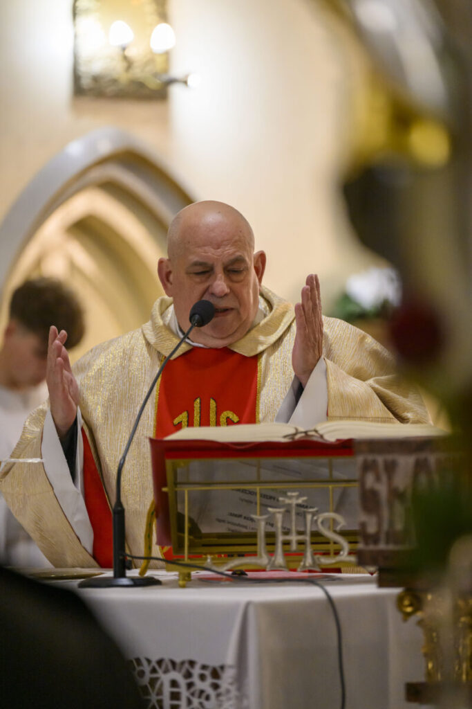 Na zdjęciu ksiądz w złotej szacie czyta przy ołtarzu. W tle widoczne są łuki i ściana w ciepłych barwach. Obok otwarta księga i mikrofon.
