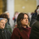 Na zdjęciu są skupieni ludzie w pomieszczeniu, przeważają stonowane kolory. Kobieta na pierwszym planie nosi bordowy płaszcz. W tle są inne osoby, ubrania w zimowej kolorystyce.