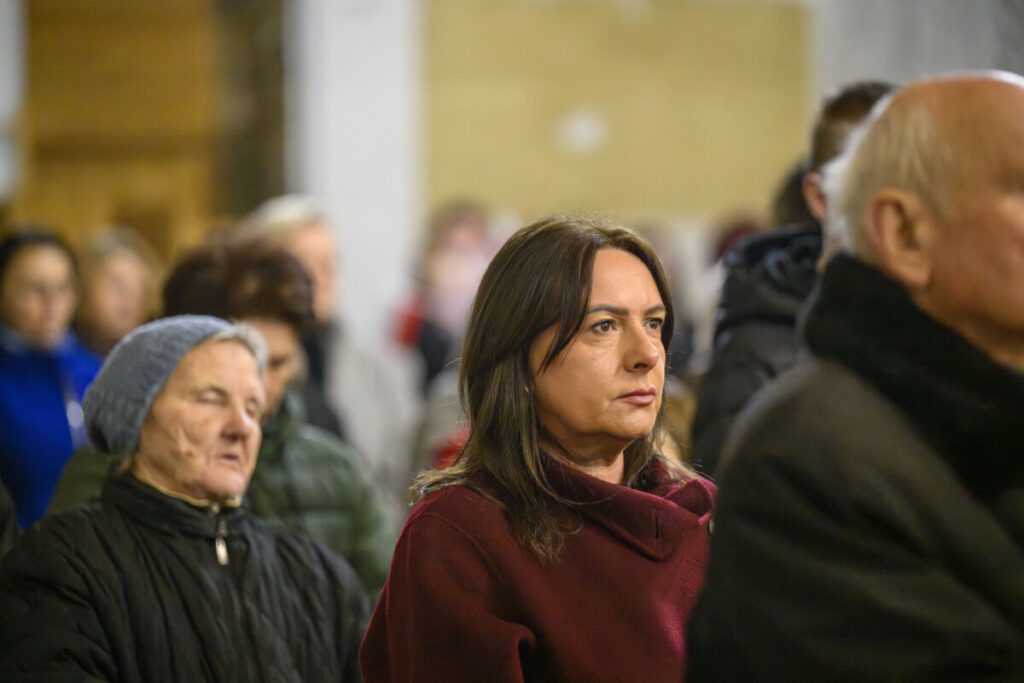 Grupa ludzi w pomieszczeniu. Kobieta w bordowym płaszczu na pierwszym planie. Rozmyte tło w ciepłych kolorach.