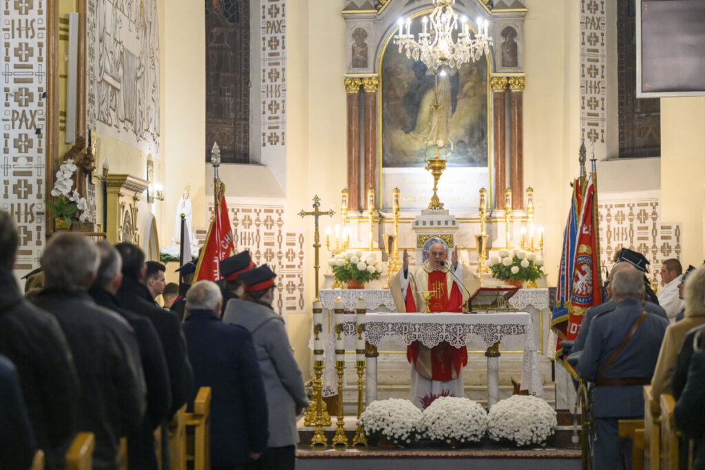 Na zdjęciu widoczna jest msza w kościele, z księdzem przy ołtarzu. W tle są jasne ściany i obrazy, wokół zgromadzeni wierni. Dekoracje w kolorach bieli i złota dominują scenę.