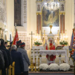 Na zdjęciu widoczna jest msza w kościele, z księdzem przy ołtarzu. W tle są jasne ściany i obrazy, wokół zgromadzeni wierni. Dekoracje w kolorach bieli i złota dominują scenę.