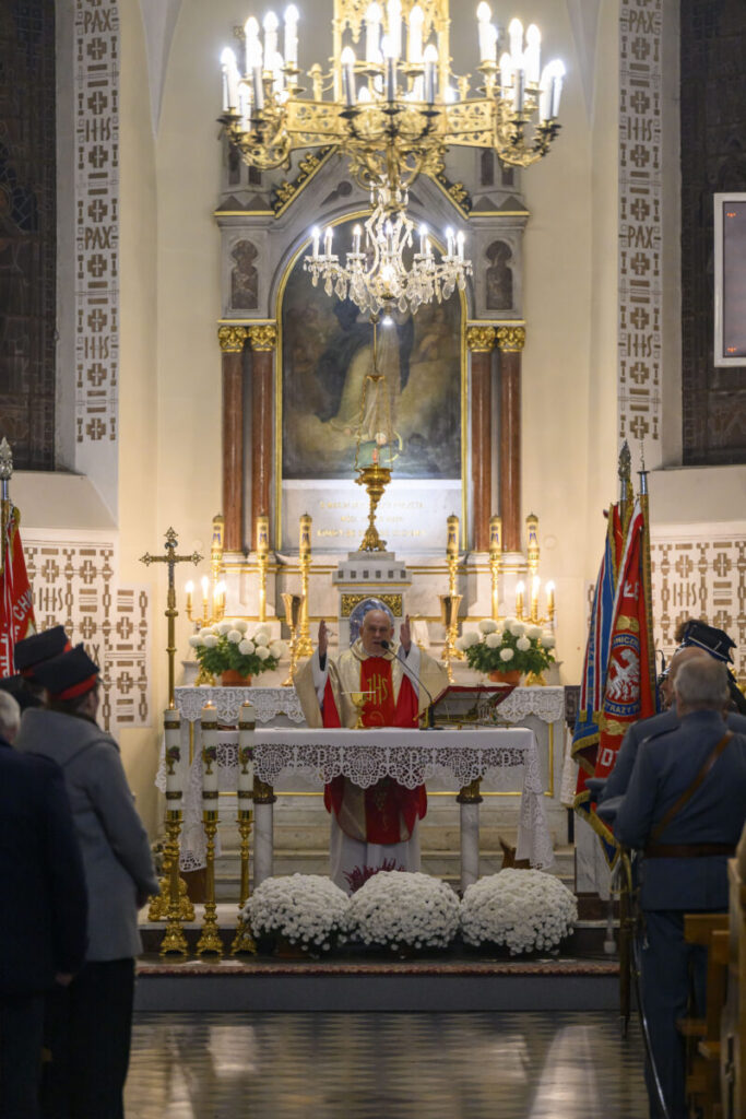 Ksiądz prowadzi mszę w kościele z jasnym ołtarzem i dużym złotym żyrandolem. Białe kwiaty zdobią ołtarz. W tle widoczne są barwne flagi i wierni.