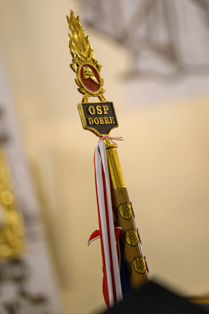 Sztandar z napisem OSP Dobre w złotych i czerwonych barwach. Tło jest rozmyte w odcieniach beżu. Przy sztandarze widoczne biało-czerwone wstęgi.