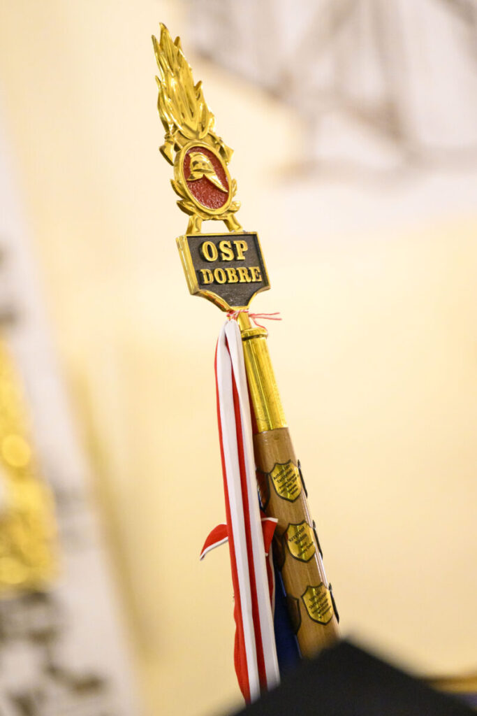 Zdjęcie przedstawia sztandar OSP z napisem OSP Dobre na złotej ozdobie. Widoczne są biało-czerwone paski w tle rozmytym. Tło jest jasne, co podkreśla detale sztandaru.