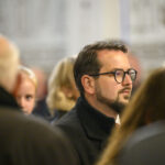 Zdjęcie przedstawia mężczyznę w okularach wśród tłumu. Dominują kolory stonowane, głównie szarości. Tło jest rozmyte, co skupia uwagę na postaci.