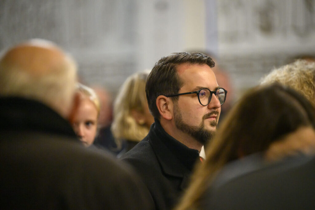 Mężczyzna w okularach patrzy w lewo na tle rozmazanych postaci. Tło jest jasne, dominują odcienie szarości. Atmosfera wydaje się poważna.