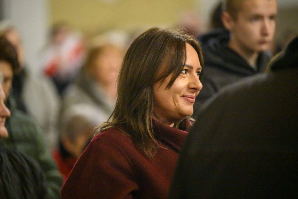 Zdjęcie przedstawia kobietę w bordowym płaszczu, z uśmiechem, w tłumie ludzi. Tło jest rozmyte, dominują ciepłe kolory.