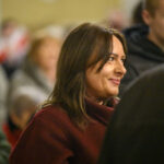 Zdjęcie przedstawia kobietę w bordowym płaszczu, z uśmiechem, w tłumie ludzi. Tło jest rozmyte, dominują ciepłe kolory.