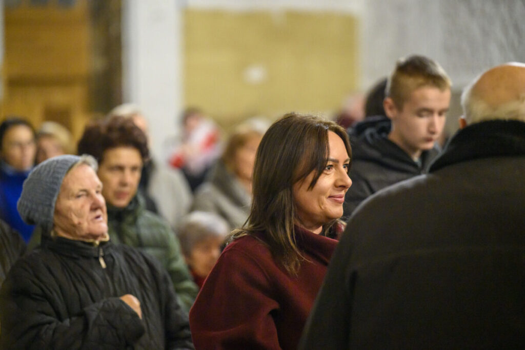 Na zdjęciu widać grupę ludzi w pomieszczeniu. Kobieta na pierwszym planie jest ubrana w bordowy płaszcz, a inni mają zimowe ubrania. Tło to rozmyta ściana w jasnych kolorach.