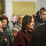 Na zdjęciu widać grupę ludzi w pomieszczeniu. Kobieta na pierwszym planie jest ubrana w bordowy płaszcz, a inni mają zimowe ubrania. Tło to rozmyta ściana w jasnych kolorach.