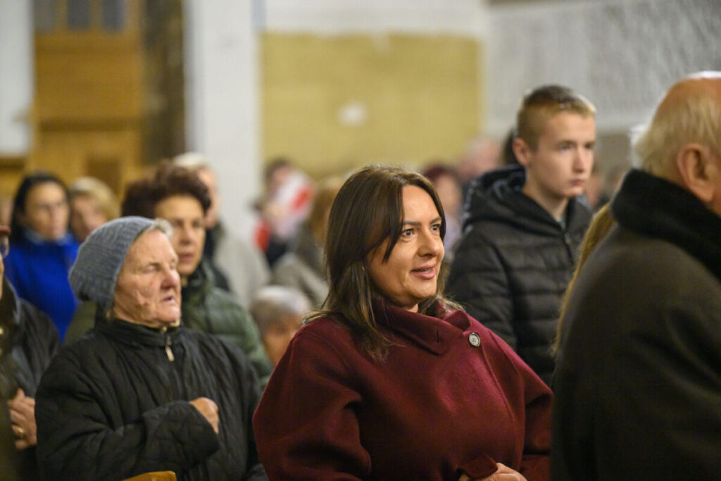 Na zdjęciu jest grupa ludzi w pomieszczeniu; kobieta na pierwszym planie ma bordowy płaszcz. Tło jest rozmyte, utrzymane w ciepłych odcieniach. Ludzie noszą zimowe ubrania.