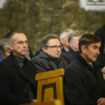 Grupa ludzi siedzi w drewnianych ławkach w kościele. Tło jest kamienne, w stonowanych barwach. Osoby wyglądają na zamyślone.