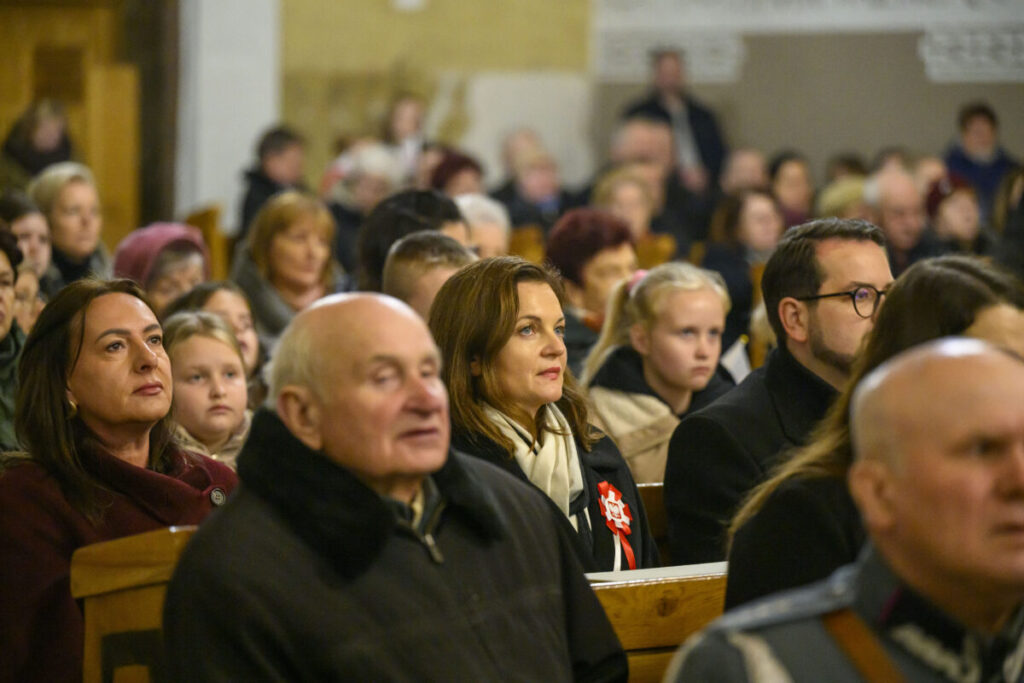 Grupa ludzi w kościelnych ławach, patrzących w tym samym kierunku. Dominują kolory beżowe i brązowe. Tło jest rozmyte, skupia uwagę na postaciach.