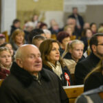 Grupa ludzi w kościelnych ławach, patrzących w tym samym kierunku. Dominują kolory beżowe i brązowe. Tło jest rozmyte, skupia uwagę na postaciach.