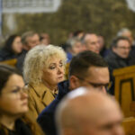 Grupa ludzi siedzi w kościele, skupiona na nieznanym wydarzeniu. Kobieta w jasnym płaszczu na pierwszym planie. Tło rozmyte, ciepłe odcienie.