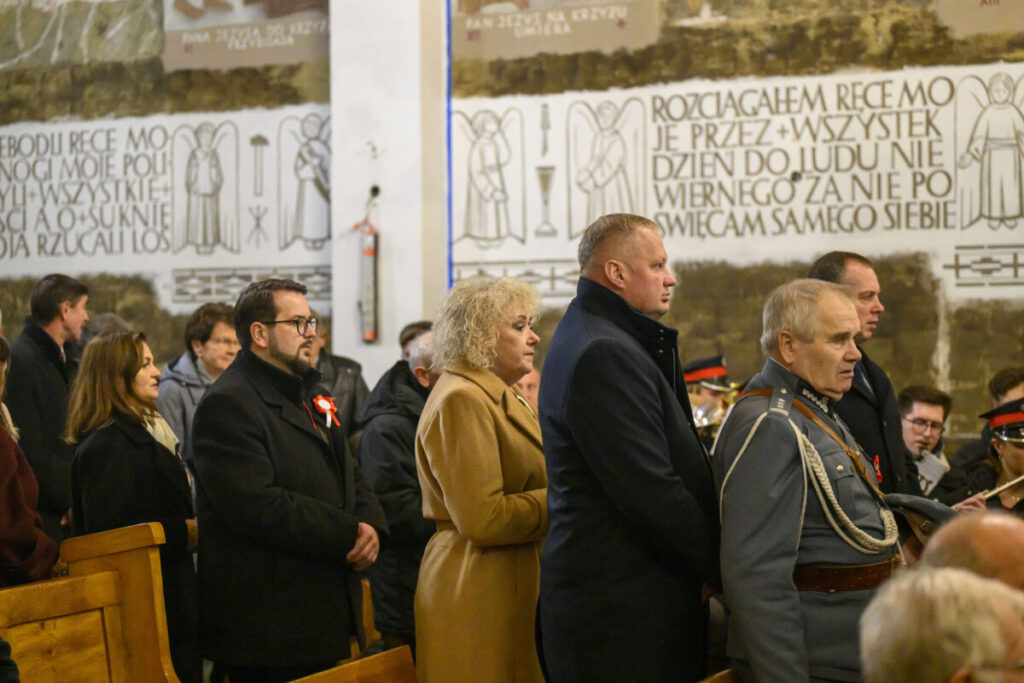 Grupa osób w kościele ubrana formalnie stoi, w tle tablica z napisem i aniołami. Dominują kolory ciemne. Po prawej stronie widać mężczyznę w mundurze.