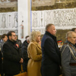 Grupa osób w kościele ubrana formalnie stoi, w tle tablica z napisem i aniołami. Dominują kolory ciemne. Po prawej stronie widać mężczyznę w mundurze.