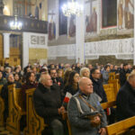 Grupa ludzi w jasnym kościele siedzi na drewnianych ławkach. Ściany ozdobione malowidłami, z białymi kolumnami. Światło żyrandola rozprasza się po całym pomieszczeniu.