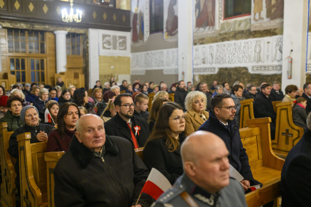 Grupa ludzi w kościele siedzi na drewnianych ławkach. Tło zdobią jasne malowidła i kolumny. Atmosfera jest poważna i skupiona.