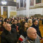 Grupa ludzi w kościele siedzi na drewnianych ławkach. Tło zdobią jasne malowidła i kolumny. Atmosfera jest poważna i skupiona.