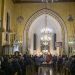 Wnętrze kościoła z licznymi wiernymi i sztandarami. Dominuje złoto, beż i czerń; sufit drewniany, tło to ołtarz. Centralnie wiszący żyrandol.