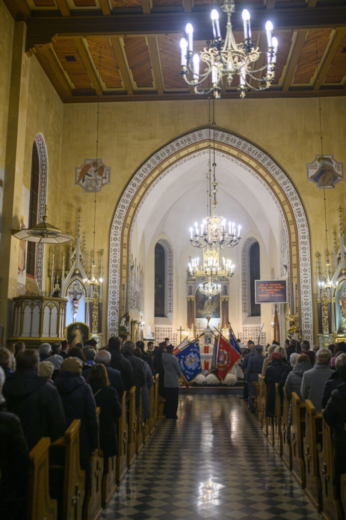 Wnętrze kościoła z licznymi wiernymi i sztandarami. Dominuje złoto, beż i czerń; sufit drewniany, tło to ołtarz. Centralnie wiszący żyrandol.