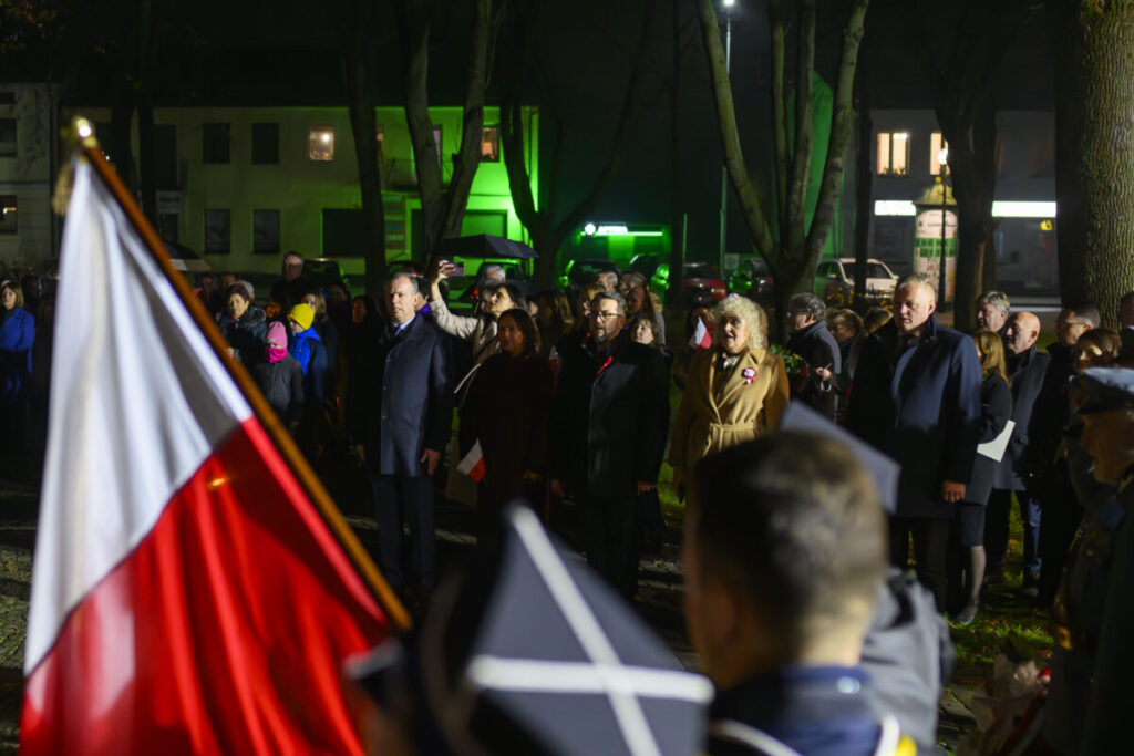 Grupa osób stoi na zewnątrz w nocy, trzymając polskie flagi. Tło oświetlone zielonym światłem, wokół drzewa. Przód zdjęcia zajmuje flaga Polski.