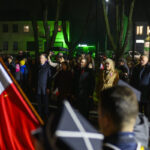 Grupa osób stoi na zewnątrz w nocy, trzymając polskie flagi. Tło oświetlone zielonym światłem, wokół drzewa. Przód zdjęcia zajmuje flaga Polski.