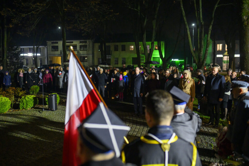 Na zdjęciu widoczna uroczystość nocą z ludźmi i flagą Polski. Tło to oświetlone budynki i drzewa. W centrum stoi grupa ludzi w ciemnych ubraniach.