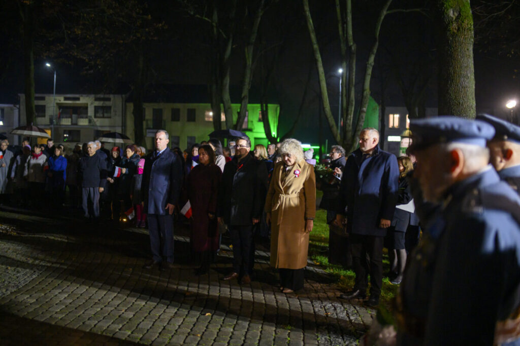 Grupa ludzi stoi na bruku, w nocnej scenerii. Tło rozświetlają zielone światła budynków. Obecne są flagi i mundury.