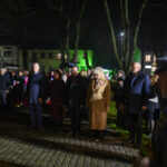 Grupa ludzi stoi na bruku, w nocnej scenerii. Tło rozświetlają zielone światła budynków. Obecne są flagi i mundury.