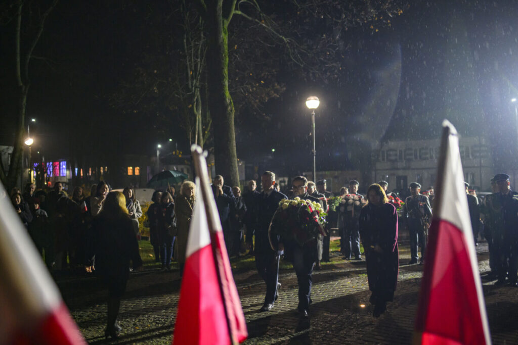 Grupa osób składa wieńce pod pomnikiem nocą. Deszcz spada delikatnie, a w tle widać flagi Polski. Jasne lampy oświetlają kamienną alejkę.