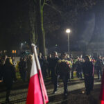 Grupa osób składa wieńce pod pomnikiem nocą. Deszcz spada delikatnie, a w tle widać flagi Polski. Jasne lampy oświetlają kamienną alejkę.