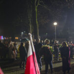Wieczorna uroczystość z tłumem ludzi i flagami Polski. Osoby niosą wieńce kwiatów, tło oświetlone latarniami. Pada lekki deszcz.