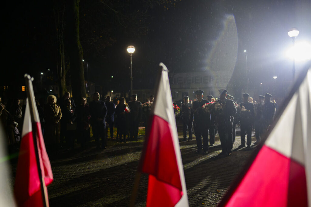 Grupa ludzi w mundurach maszeruje nocą, trzymając kwiaty. Na pierwszym planie widoczne są flagi Polski. W tle lampa uliczna oświetla ciemne otoczenie.