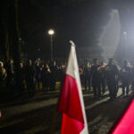 Grupa ludzi w mundurach maszeruje nocą, trzymając kwiaty. Na pierwszym planie widoczne są flagi Polski. W tle lampa uliczna oświetla ciemne otoczenie.